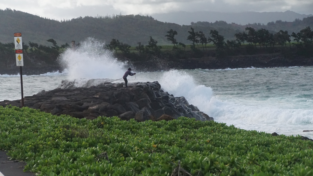A man stands tries to bait a hook attached to a long fishing pole while standing on an artificial stone embankment as waves crash into the stones sending spray well above his head