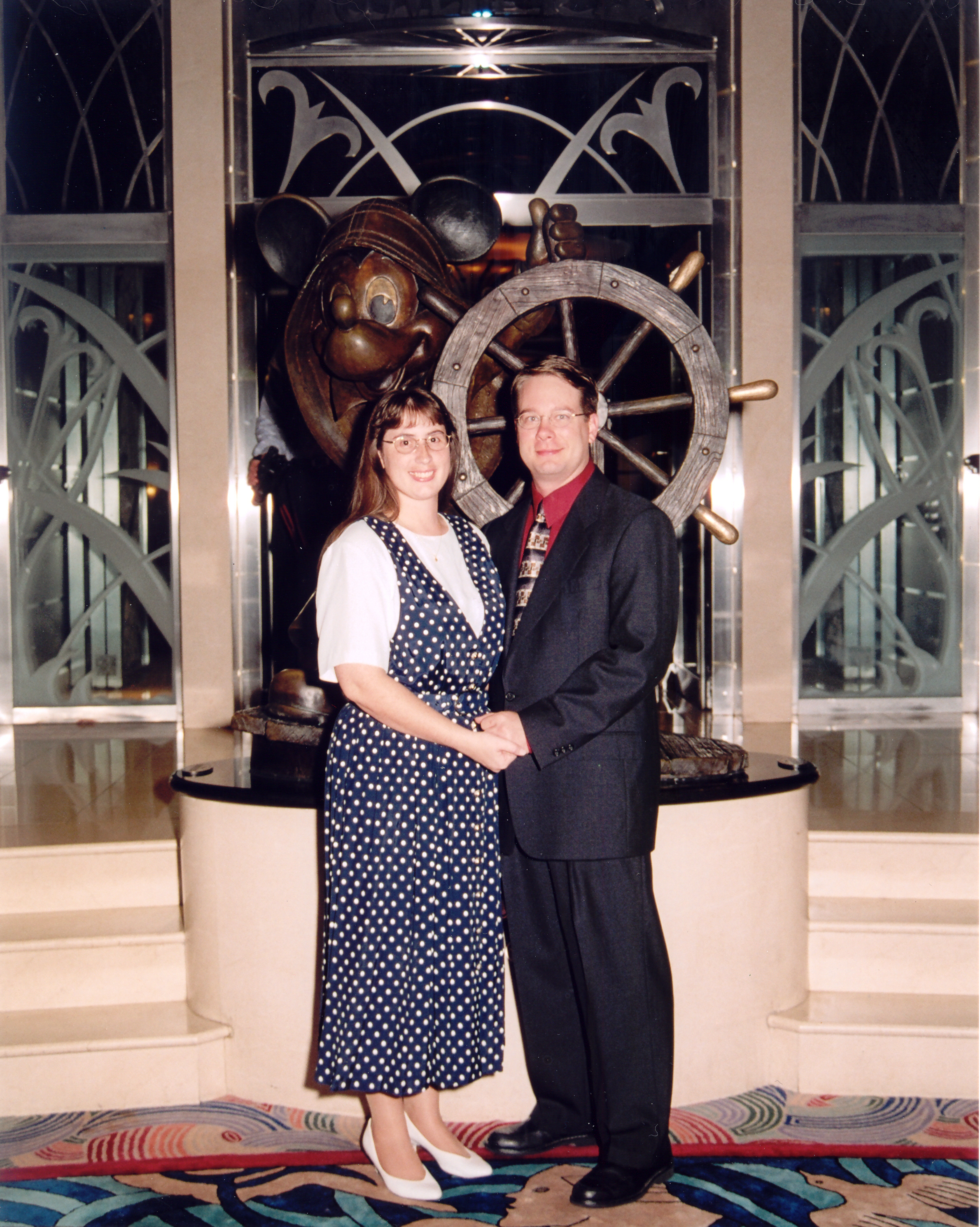 James and Julie dressed up in front of a statue of Mickey Mouse at a ship's wheel