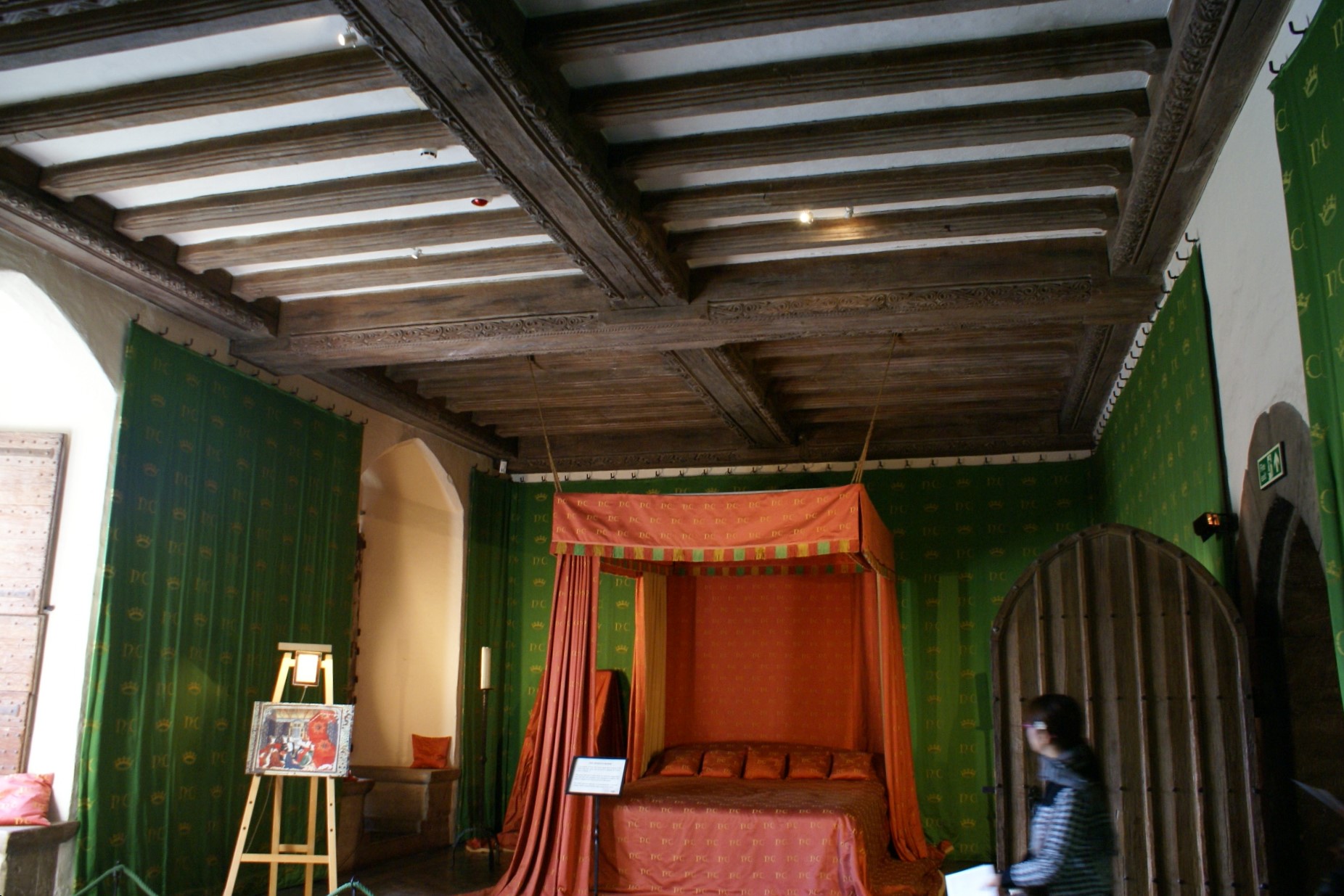A bed with red surround in a room with green tapestry walls and wood beam ceiling