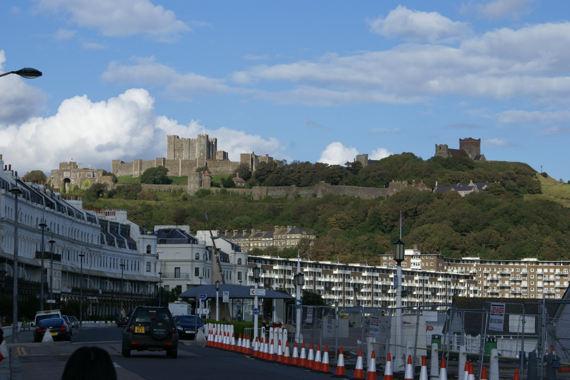 An expansive castle on a hill overlooks modern buildings along a curved road along the water
