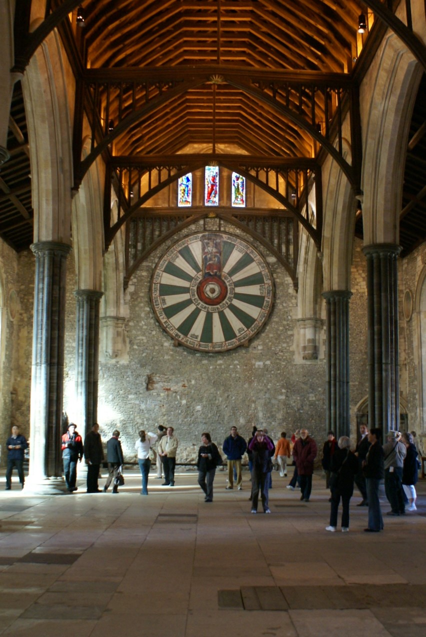 A large round tabletop with a king and 7 names on green and white sections hangs on the wall below a wood beam roof structure on then stone columns