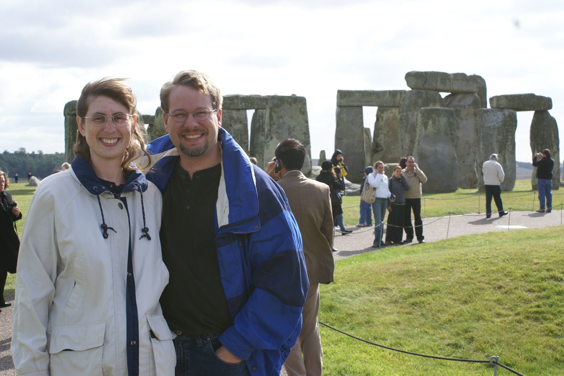 James and Julie stand on a path with the standing stones of Stonehenge beyond