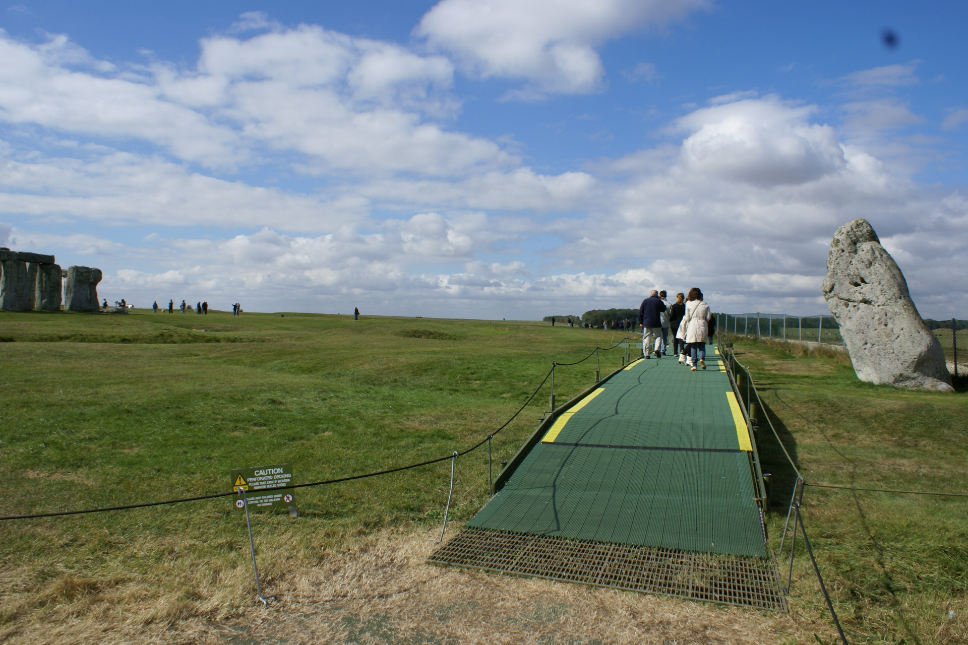 A single rough stone stands near the walking path a distance away from the standing stones