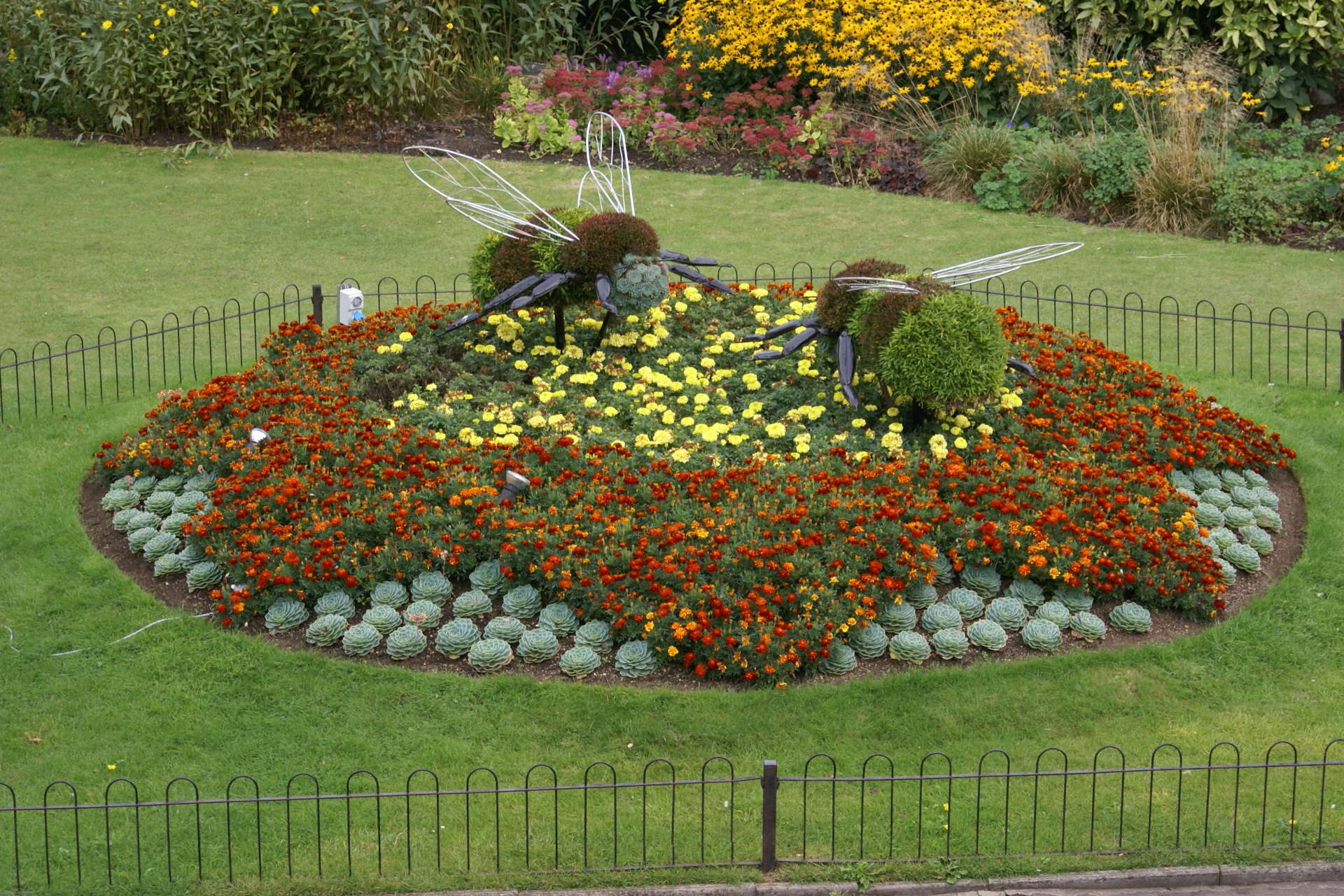 A flower bed with orange flowers in a star shape and yellow center has two bumble bee topiaries