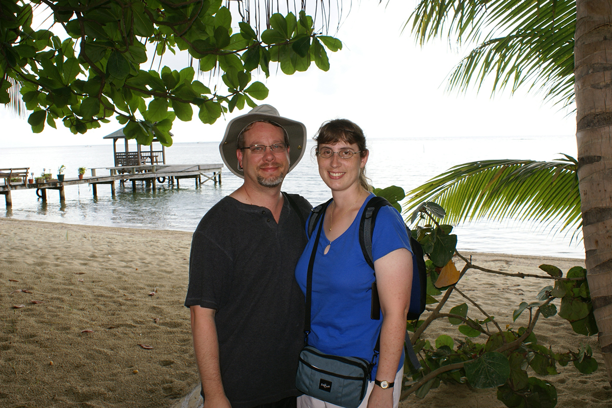 James and Julie under a tree branch with a wood dock running into the ocean beyond