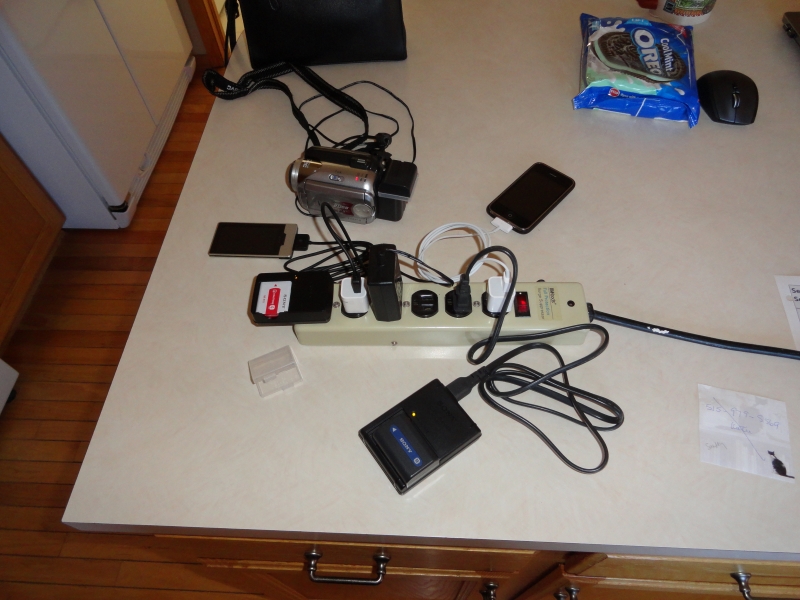 A power strip on a counter is filled with charging cables to cameras, battery packs, phones, and audio players