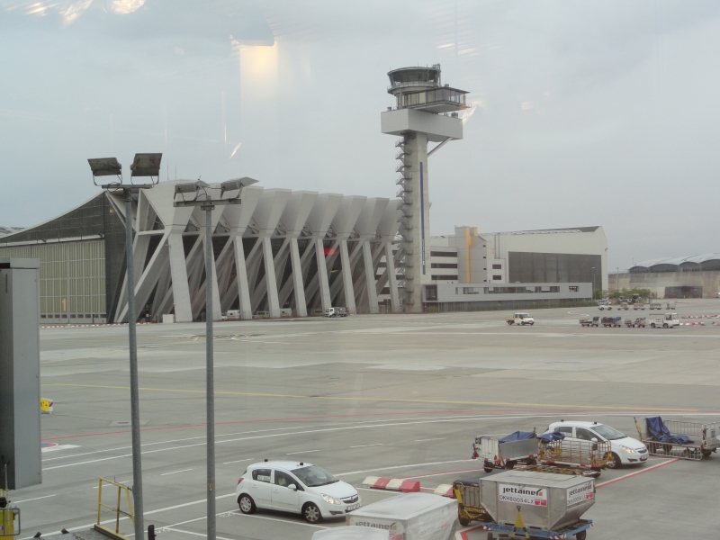 A concrete tower with round glass space at the top extends high above sweeping concrete forms supporting a roof at the edge of a large concrete area where airplanes can maneuver