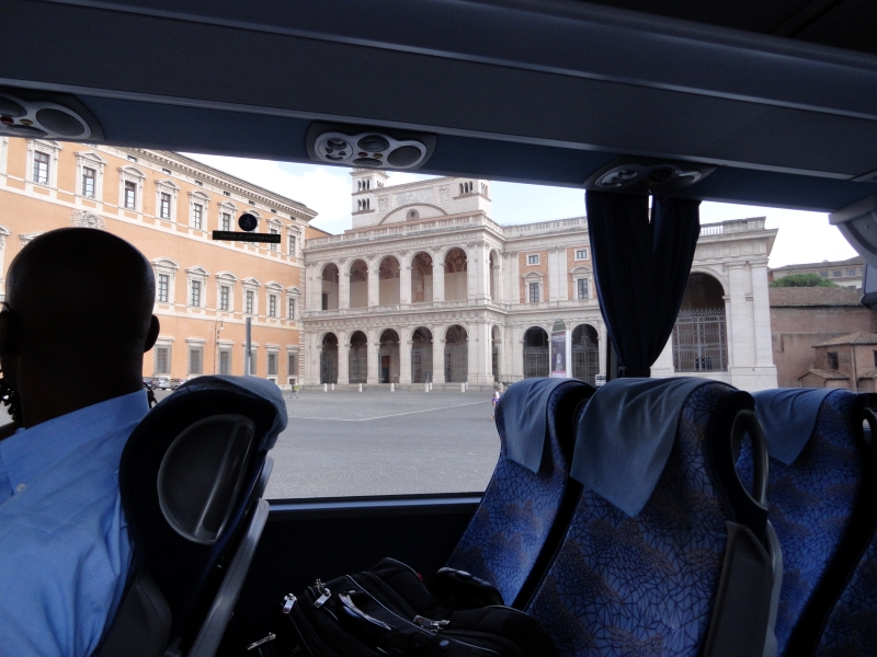 Looking out the coach window at a plaza of black stone pavers in front of a two-story white stone building with arches and ornamental gates attached to a three-story tan building with ornamental stone window surrounds