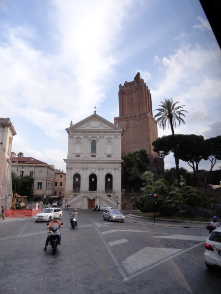 A white church with steps and three arched openings stands off a traffic circle with the ruins of a much taller red brick tower beyond