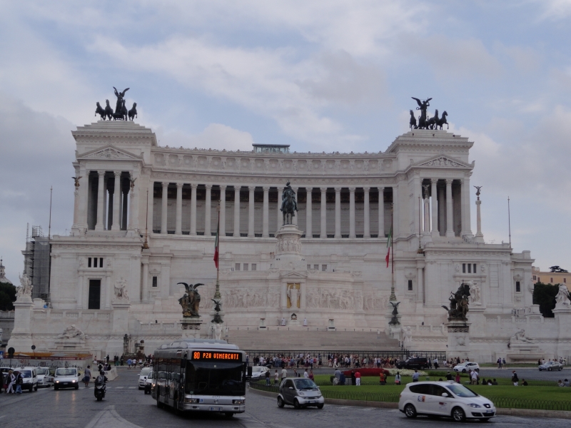 A massive bright white building with many carvings is surrounded with large bronze statues and stands beyond a busy traffic circle. The building has huge columns like an ancient temple above a multistory base with very few openings.