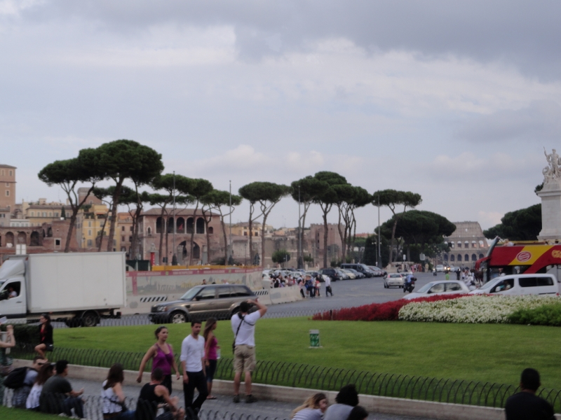 A large parking area near historic buildings of various colors of stone standing beyond tall trees with bushy green tops. In the distance the walls of Rome’s Colosseum are visible