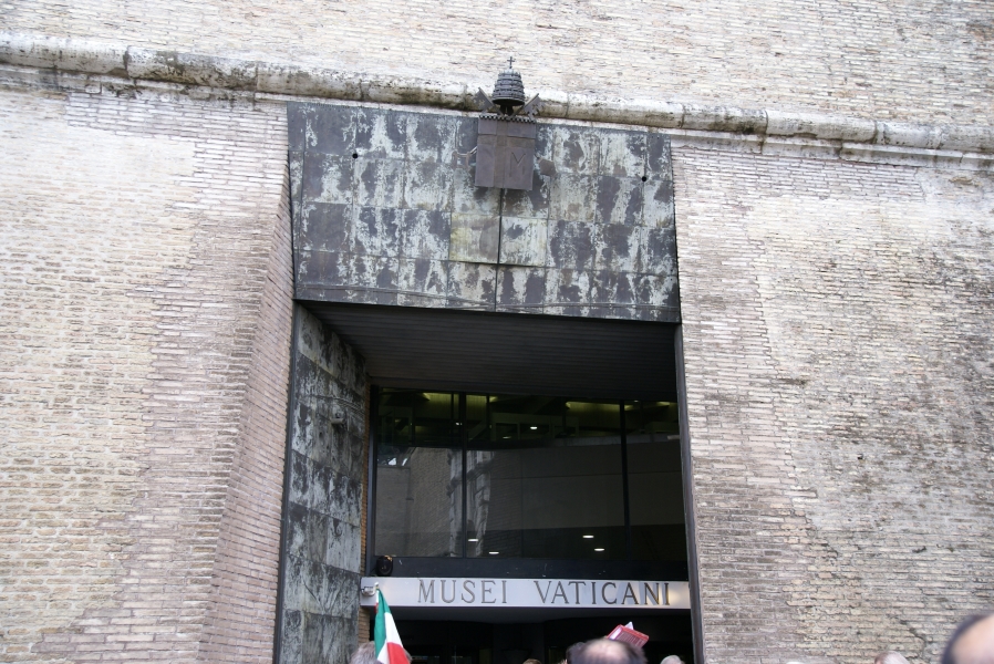 An opening in a white brick wall has glass wall with a sign reading Musei Vaticani below an aged steel header with a three-dimensional crest of the Vatican
