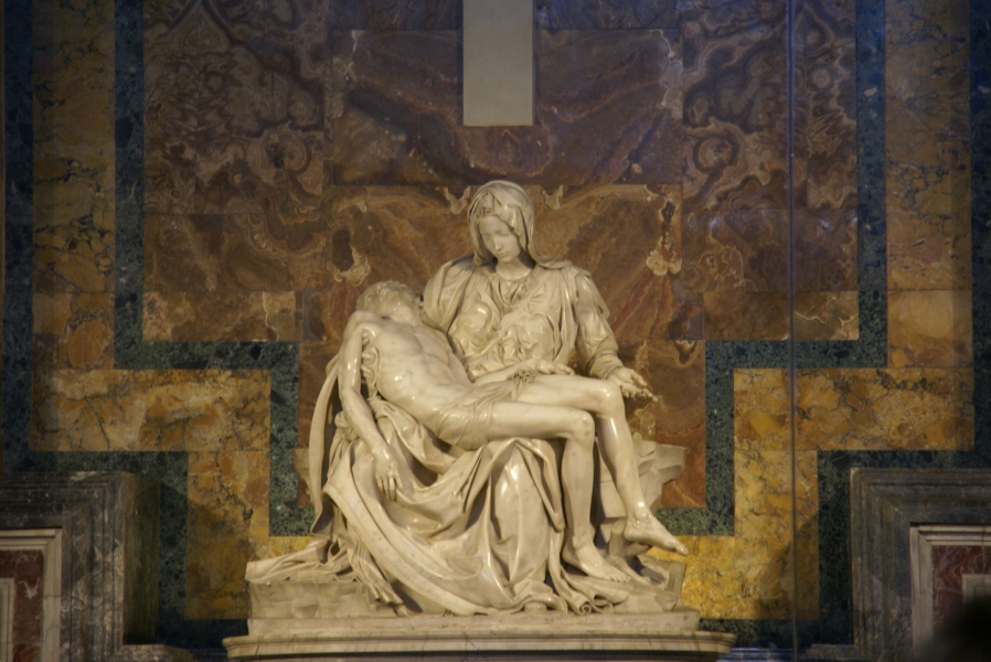 A brownish tan marble statue shows a very lifelike woman holding the body of a deceased man in her lap
