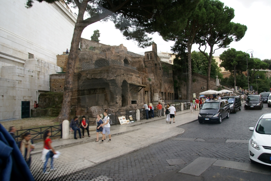 Ancient reddish stone and brick ruins fill a wedge between a sidewalk, stair, and modern white stone building below towering trees