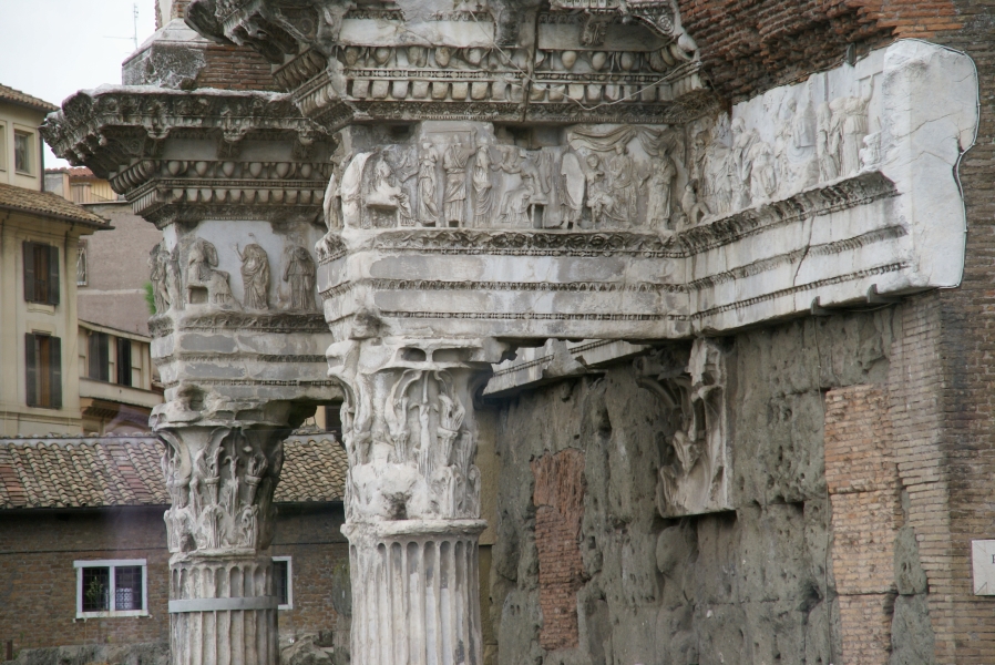 Weathered carvings adorn white stone bands and columns sticking out from the face of a red brick building with concrete filling in portions of the wall to keep everything stable