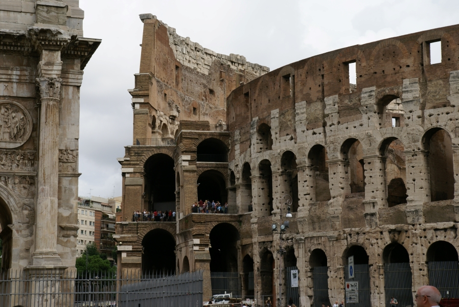 Three concentric curved walls of archways are connected with other arches where people stand on a second level thirty feet above the ground. The walls are a combination of white weathered stone and red brick