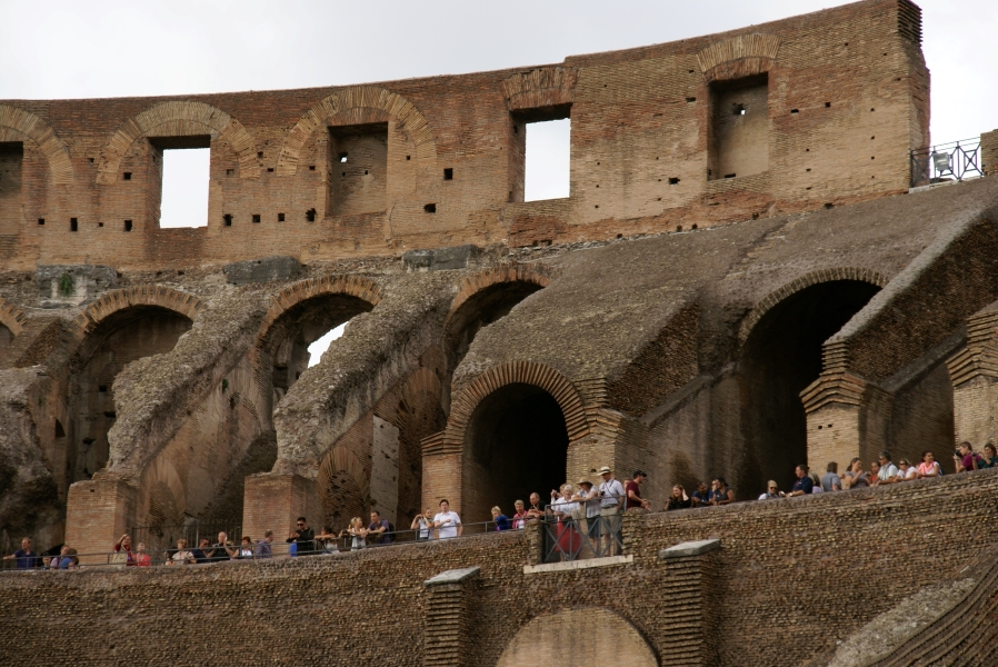 People line the railing of an elevated walkway with large, sloped walls over arches behind them