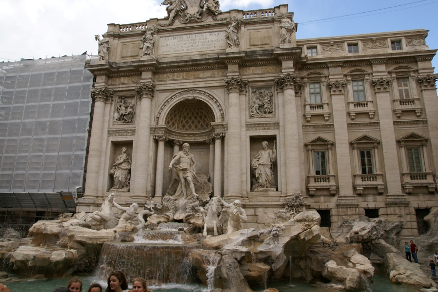 A white stone façade has a large pool in front above which carved stone figures work to contain horse-like sea creatures as water flows amongst a carved landscape