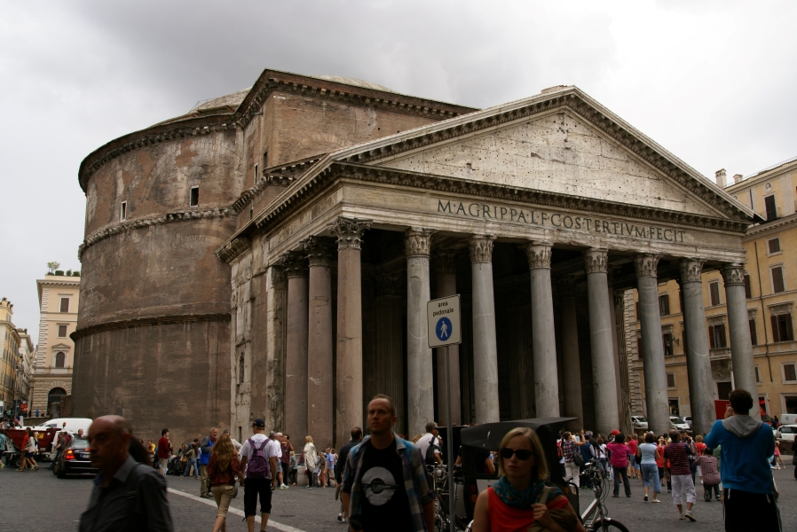 A four-story reddish brown round building with a classical white stone front portico with 20-foot-tall columns with carved capitals sits surrounded by blocky buildings