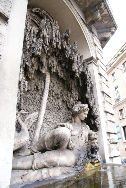 A small fountain nestled into a corner of a building has a figure reclining under a tree with a swan flapping its wings