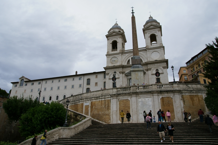 Wide steps lead to a landing where they split and rise to another level where a white stone church with two bell towers stands behind a reddish-brown obelisk with a fleur de lei and a cross on top