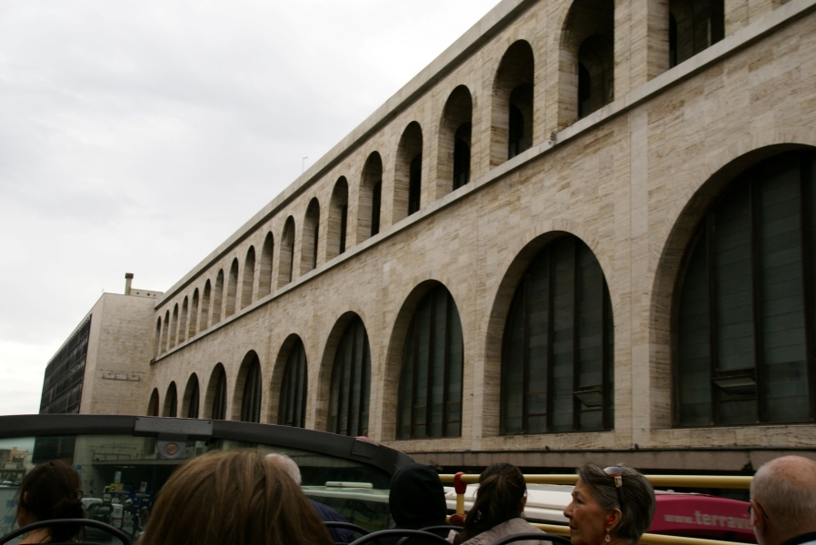 A five-story wall of white stone with two levels of different size arches above a street level canopy runs along a street where buses are parked along the curb