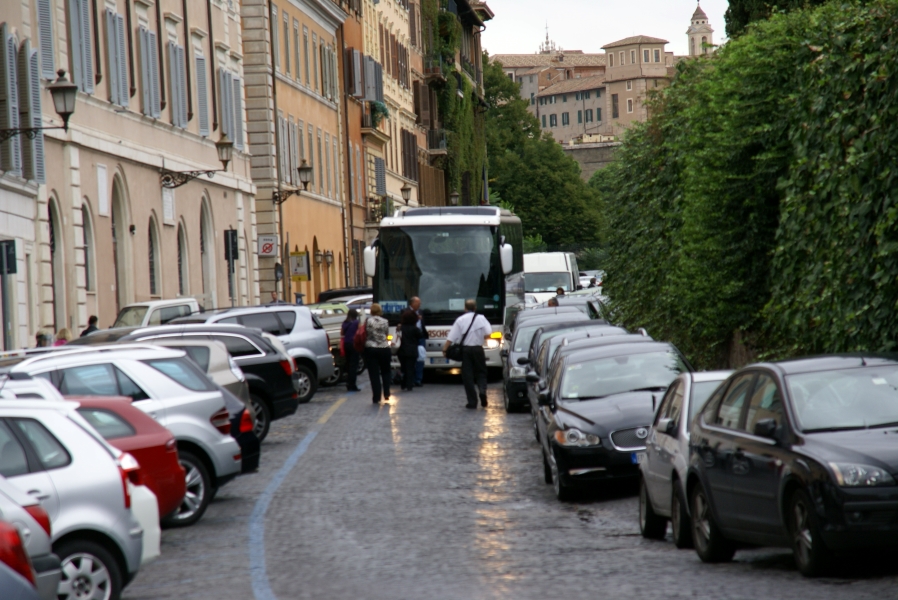Several people stand in the street with cars lined up on both sides in front of a passenger coach with a line of cars behind it