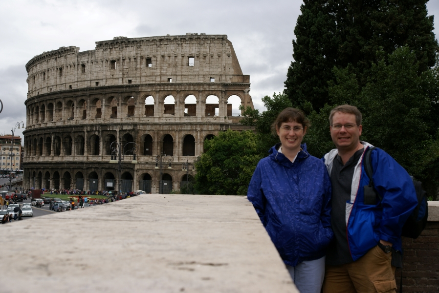 Julie and James stand next to a chest high stone capped wall with green trees behind them and the ruins of a stone building with three levels of arches and a solid wall in total standing over a 100-feet tall is in the distance