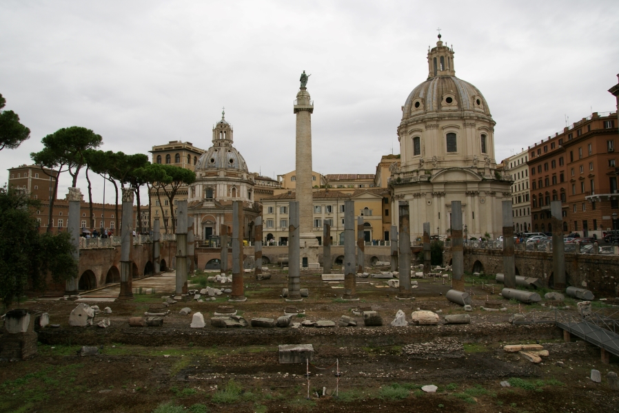 Two rows of stone columns run through a field of grass and stone rubble between two arched walls supporting modern sidewalks and streets with a taller white stone column with a statue on top