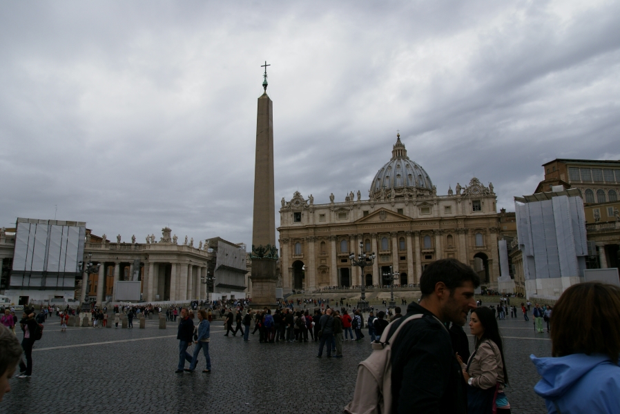 Groups of people stand in a large plaza around a tone obelisk in front of a white stone church façade with curving arched bands extending from the steps to the church encompassing both sides of the plaza