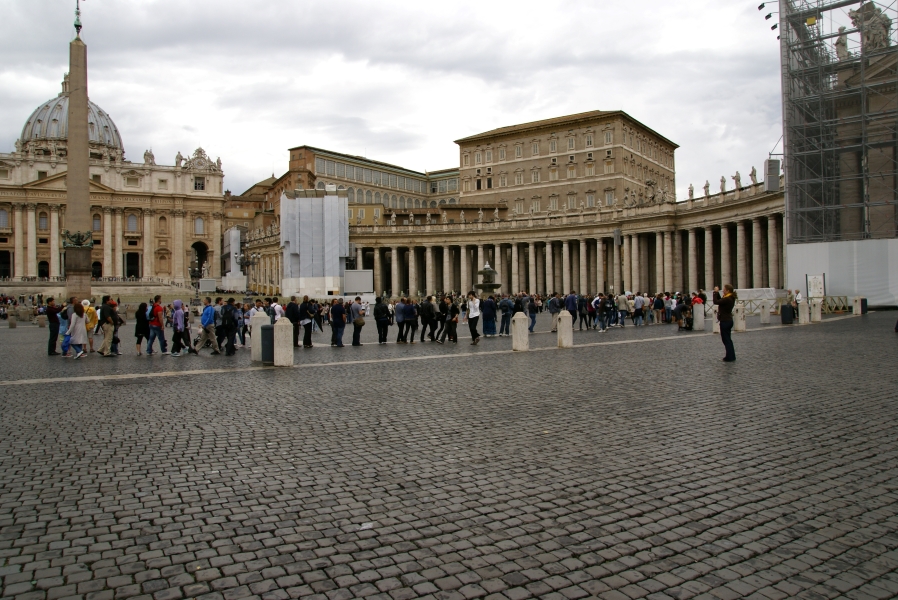 A stone church entrance on the left edge of the photo turns into a curved colonnade extending around a plaza where around 400 people stand in a line to pass through security and enter the basilica