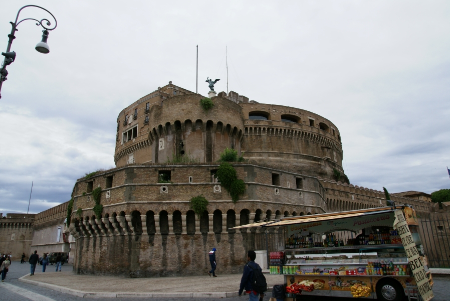 A food truck with fresh fruit and snacks stands in front of a large red brick structure with multiple curved forms of increasing height has they get closer to the center where a large patinaed green angel statue stands above the whole structure