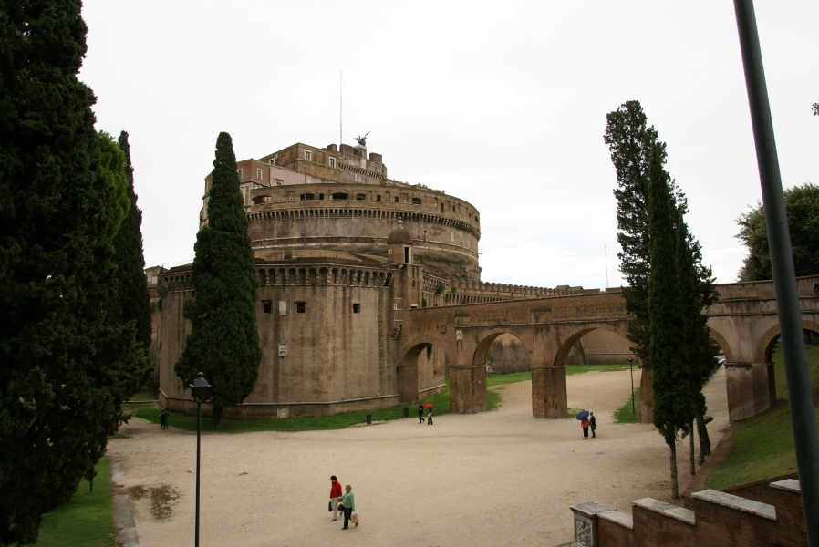 A narrow-arched stone elevated path crosses gravel paths acting as a dry moat against tall walls in front of a large round structure