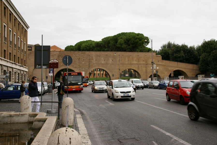 A red brick enclosed, elevated walkway supported by large arches crosses a major road before sloping down and disappearing into a stand of trees