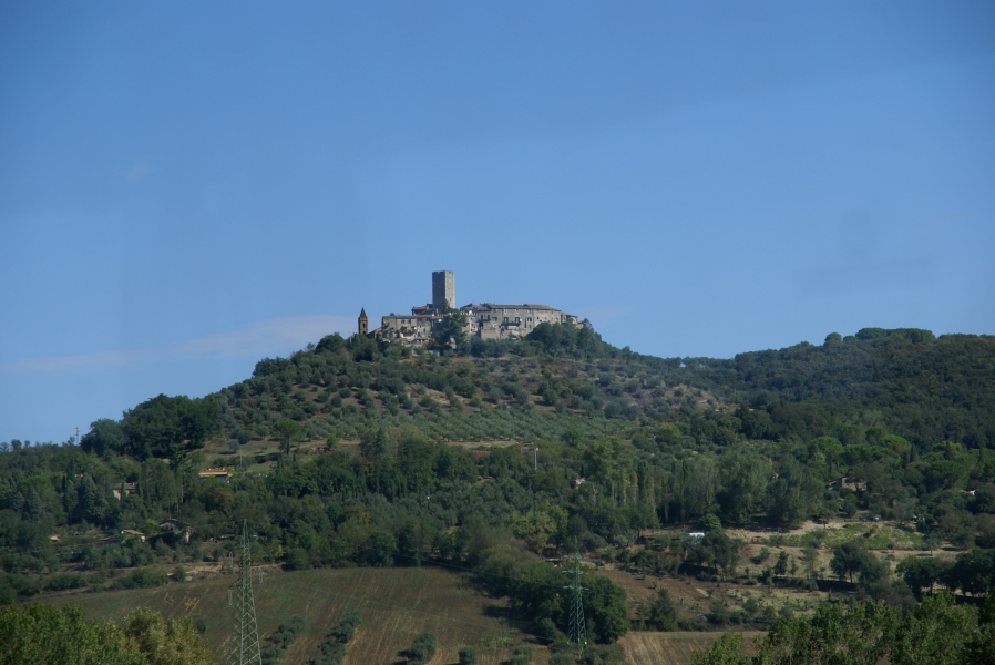 A square tower of gray stone stands above a walled city on a hilltop overlooking olive tree groves