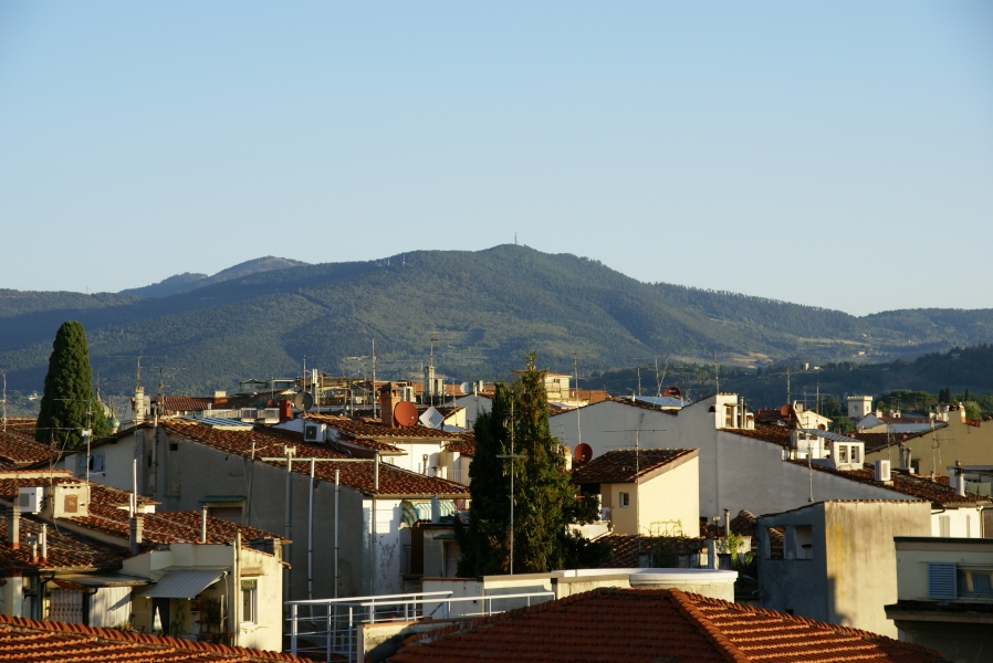 White stucco buildings with sloped red tile roofs fille a valley below green tree covered hills with the early morning sun shining down