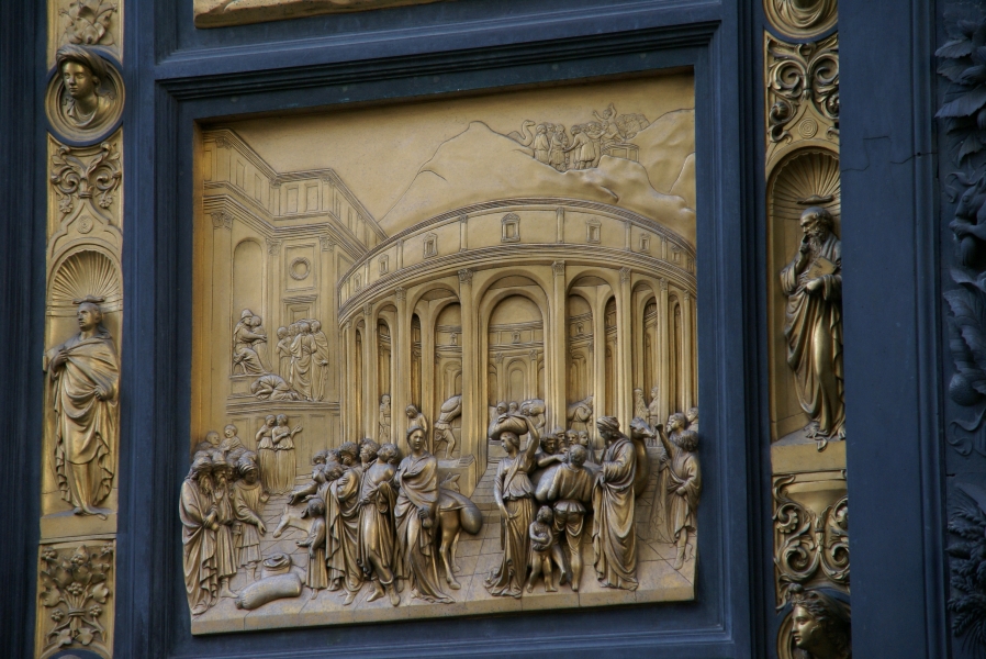Bronze bands of detailed reliefs run outside a black frame surrounding another bronze relief depicting people carrying various food goods outside a circular structure while others stand atop mountains in the background