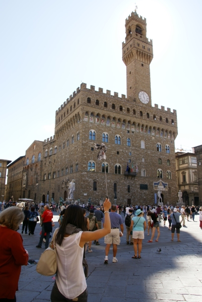 A brown stone block 5-story structure with a clock and bell tower extending another 5 stories sits on the edge of an open area filled with large statues