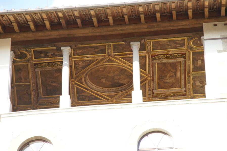 Intricately painted ceilings with gold trim can be seen through openings in an exterior wall along an high walkway