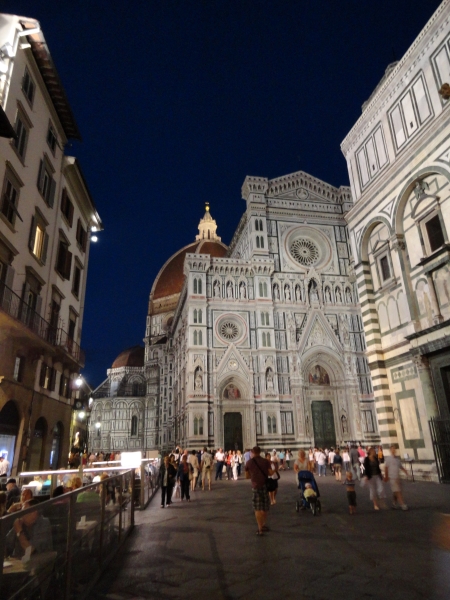 Lights illuminate the tile mosaics on the front of the Florence Cathedral with the Dome cap brightly lit against a dark sky as people sit in cafes along the edges of the piazza in the evening