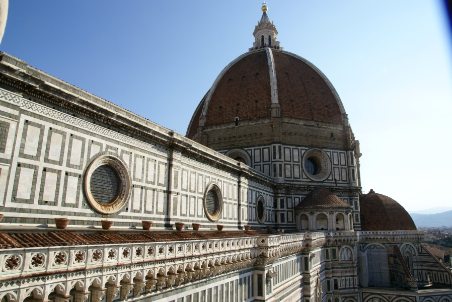 A wall of white stone tiles with green rectangular accents and round windows runs along the below the upper roof to a much taller octagonal structure with similar materials supporting a ribbed dome of red terra cotta tiles with a white couple and cold cross at the top