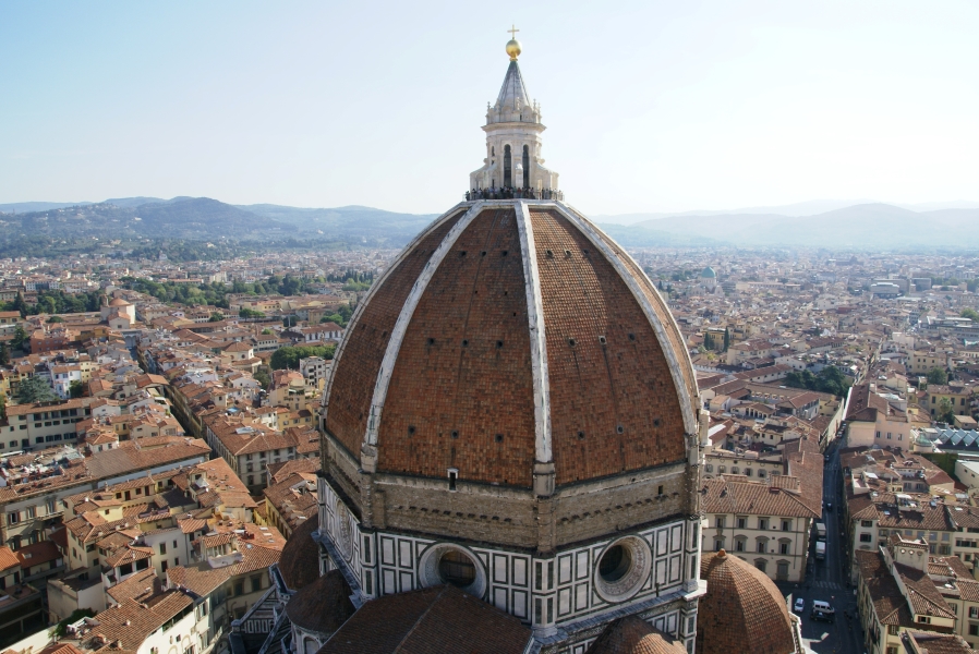 Red tile roofs of buildings spread into the distant haze where shadows of green hills are visible behind the white ribs and terra cotta tiles of the Florence Cathedral’s dome where people standing at a railing around a white structure and cold ball and cross top