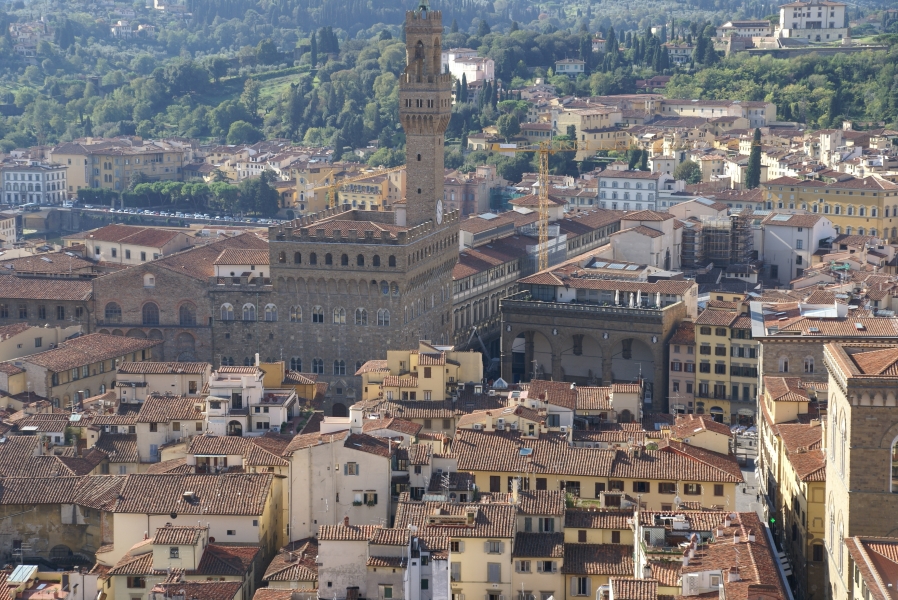 Looking out and slightly down on the brown stone block clock and bell tower of Palazzo Vecchio raises above the other buildings nearby including the café of the Uffizi Museum