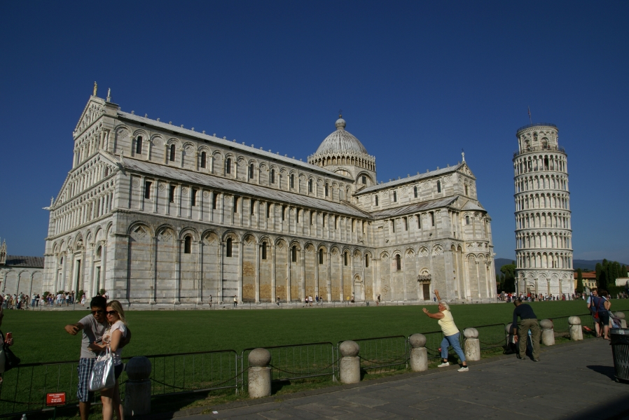 People stand along a railing taking pictures of the green lawn and large white stone Romanesque church with free standing circular bell tower beyond; the bell tower is obviously leaning to one side