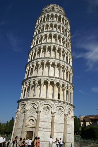 A round tower of white stone with seven levels of arched rings and a stone block base sets behind a railing and the door at the bottom shows the building is leaning far to the right