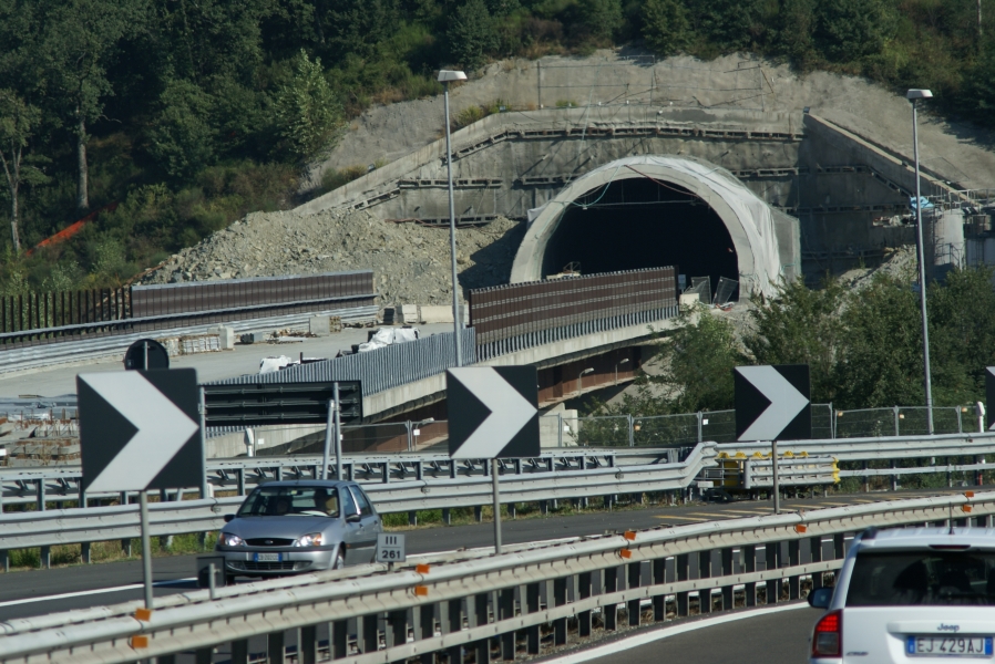 A bridge sized for multiple lanes of vehicle traffic runs into the mouth of a tunnel under construction pierces the side of a hill