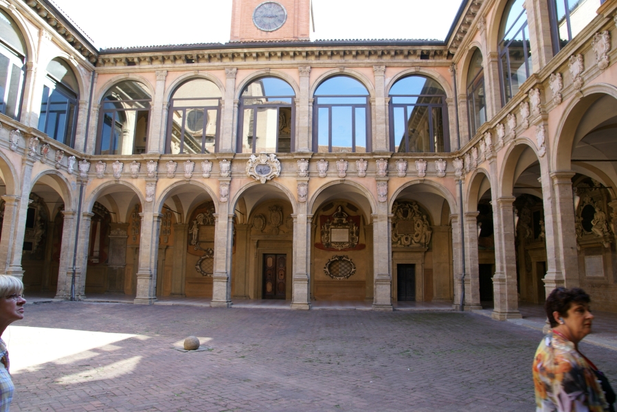 A courtyard of red brick pavers is surrounded by a story structure with arched openings enclosed with glass on the second level creating a covered walkway on the ground level with large decorative plaques