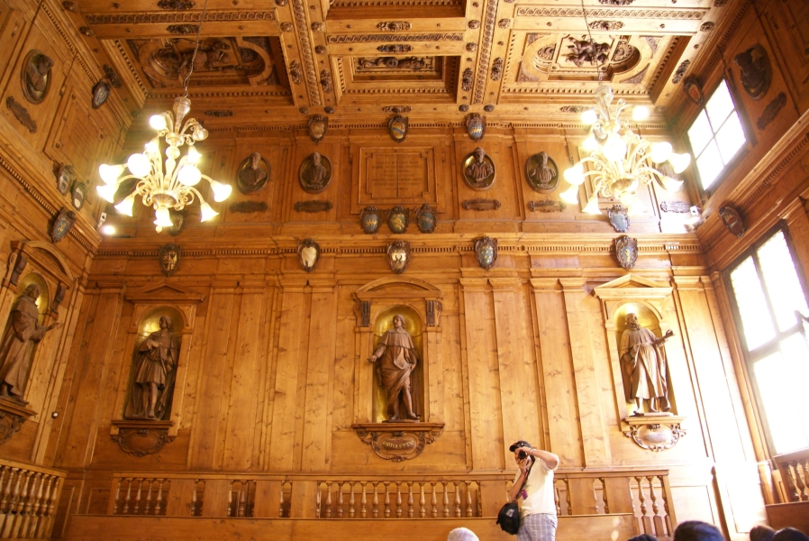 Ornate wood carved trim, statues, shields, and busts fill the walls and ceilings of a wood panel lecture hall with elevated walkways and railings around the perimeter of the room