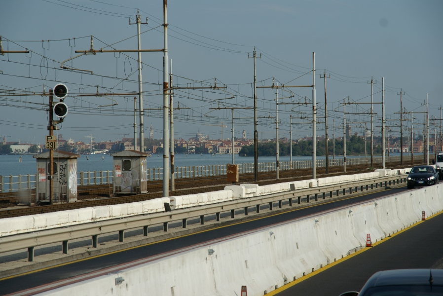 Bell towers and domes extend above buildings set low to the water are visible beyond rows of power lines used for trains crossing the bridge they share with car and coach traffic to the depots serving Venice