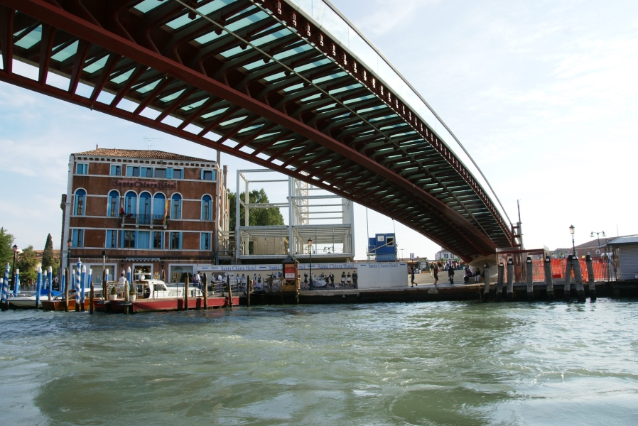 A curved steel frame and ribs supports a glass walking surface and railing for a bridge crossing a canal of greenish blue water